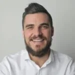Portrait of a young bearded man smiling, wearing a white shirt indoors against a gray background.